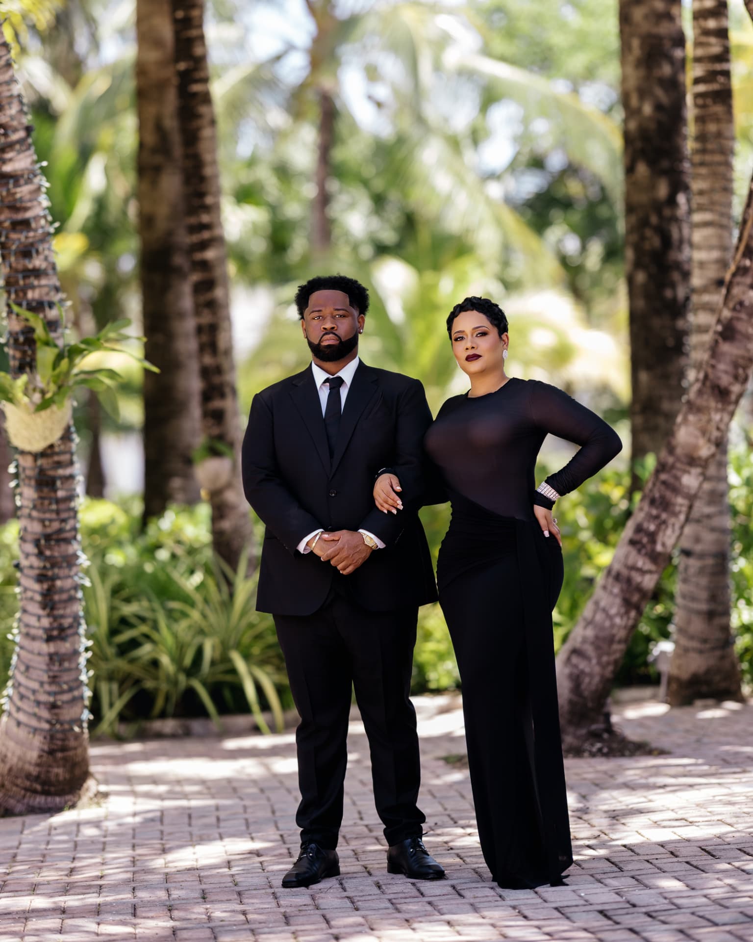 Lathario in a black suit and tie and Drew in a floor-length black gown, standing arm in arm among palms